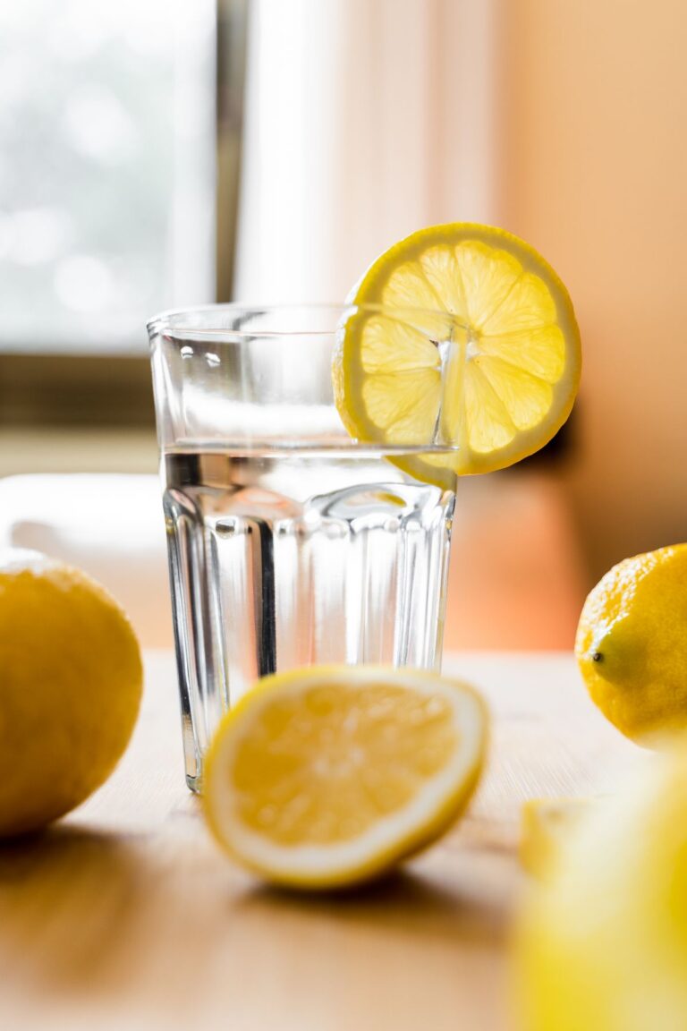 Glass of lemon water with a lemon wheel on the rim, whole and halved lemons beside it on a wooden table in bright light