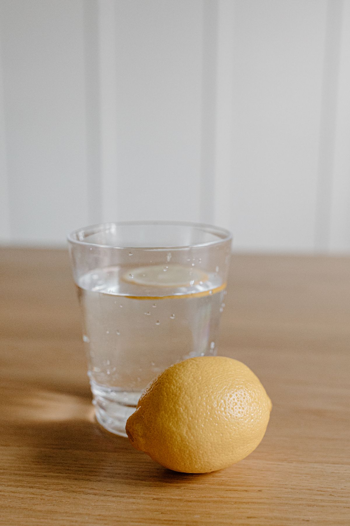 Glass of lemon water with a lemon slice and a whole lemon on a wooden table in soft morning light