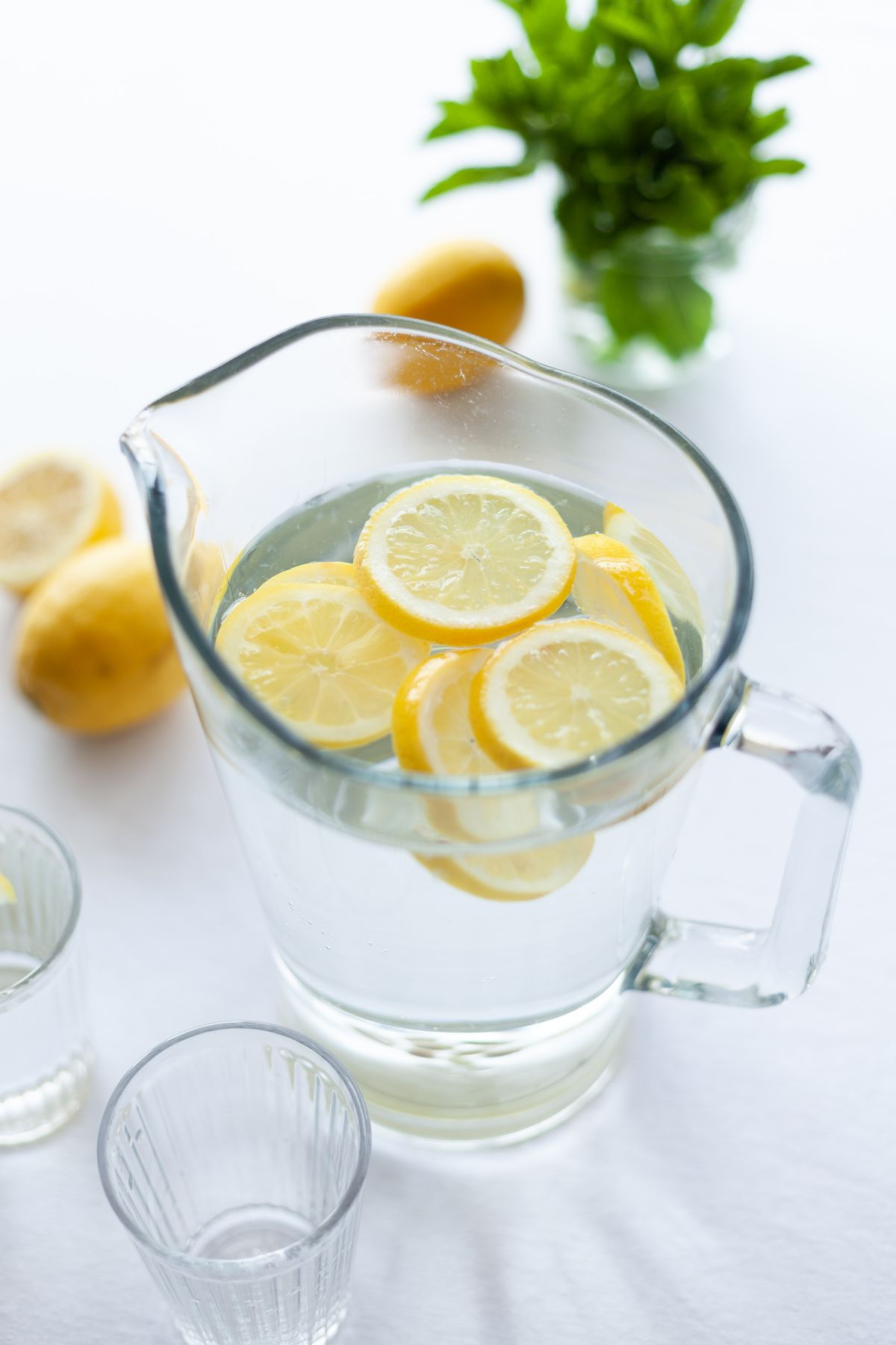 Pitcher of lemon water with lemon slices, drinking glasses and fresh mint on a white table